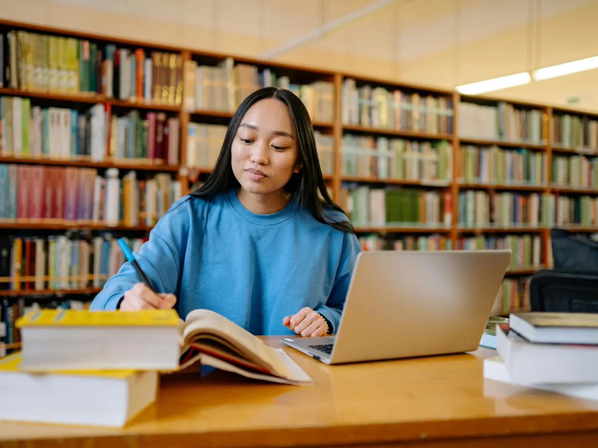 A student writing notes beside an open book and a laptop in a library