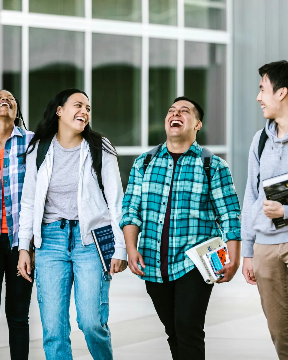 Four students with backpacks and books walking and laughing outside a university building